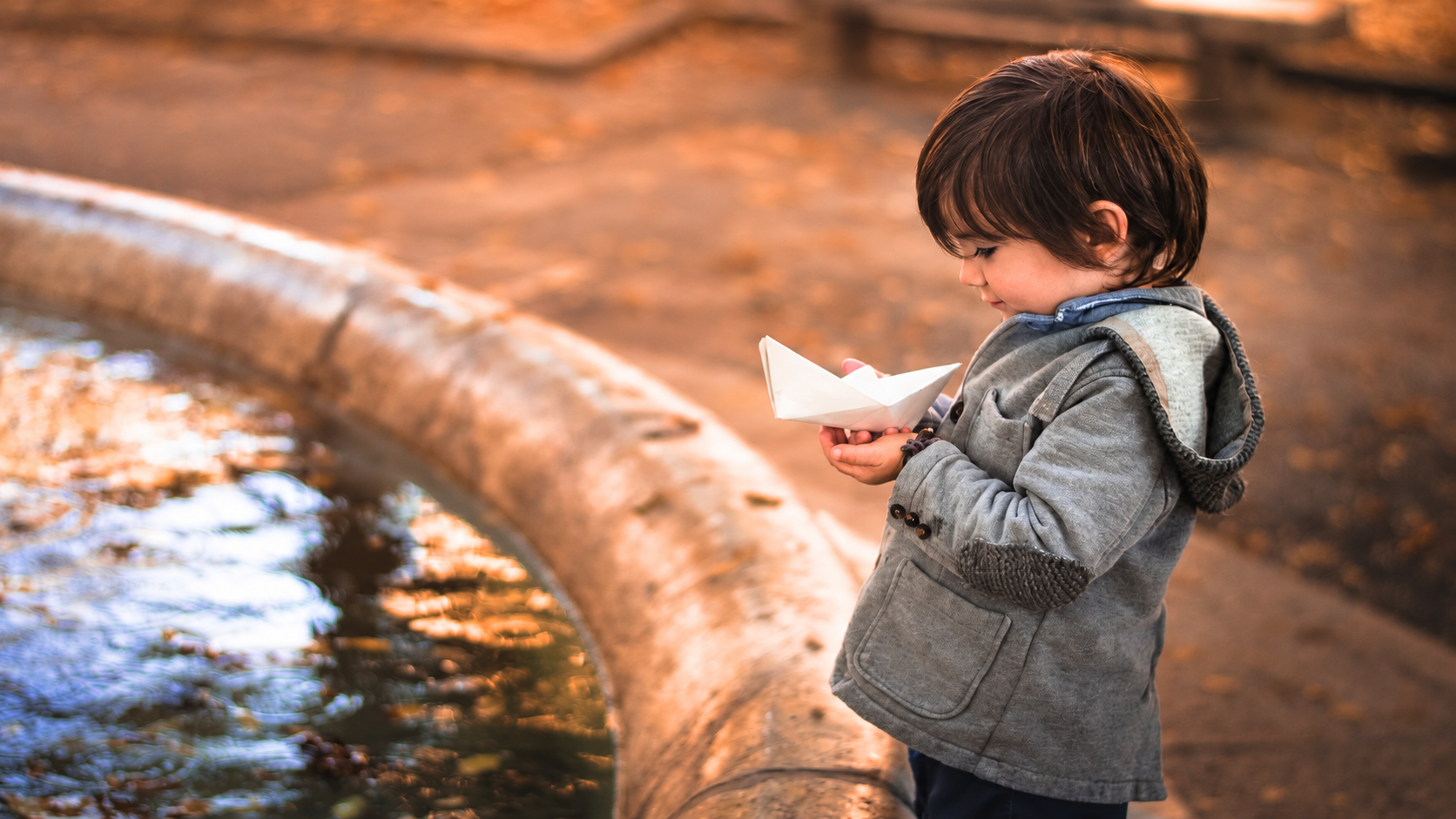 Un bambino osserva la sua barchetta di carta accanto all’acqua.